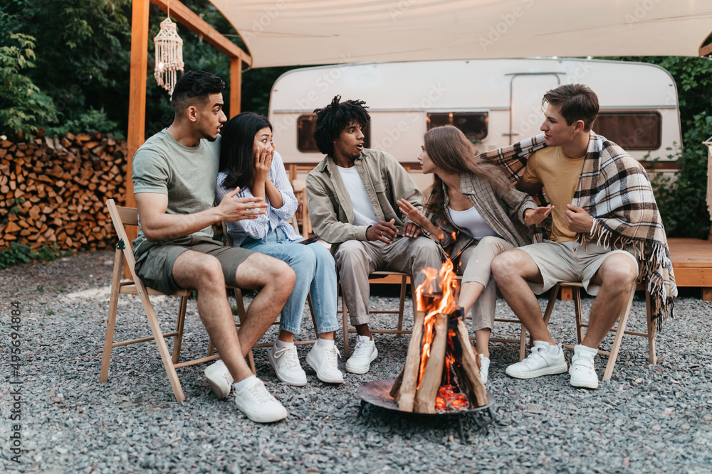 Diverse young friends sitting near campfire, telling scary stories next to RV during camping trip on summer vacation