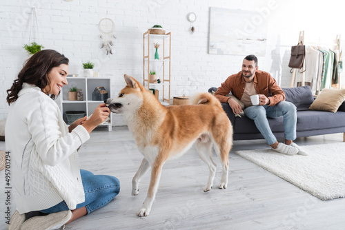 happy young woman playing with akita inu dog near bearded boyfriend with cup of coffee.