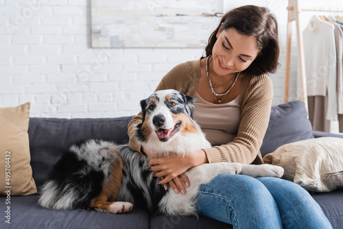 happy young woman cuddling australian shepherd dog while sitting on couch.