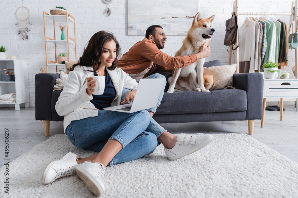 young woman with cup sitting on carpet and using laptop near boyfriend cuddling akita inu dog. young woman with cup sitting on carpet and using laptop near boyfriend cuddling akita inu dog.