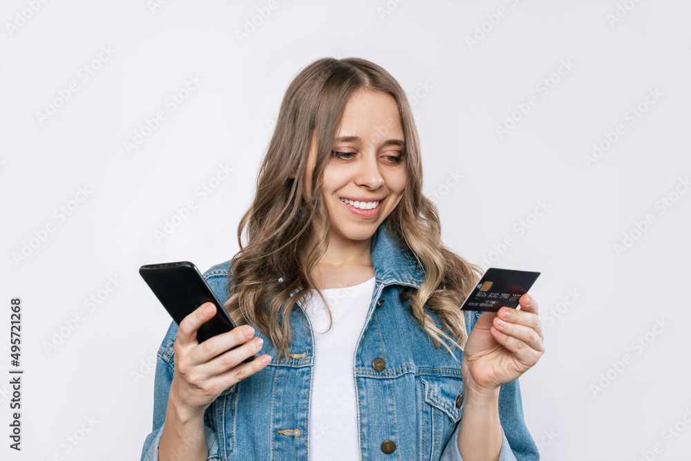 A young charming blonde woman with wavy hair in a denim jacket holds a plastic credit card in her hand looking at the mobile phone screen while making an online purchase isolated on a white background A young charming blonde woman with wavy hair in a denim jacket holds a plastic credit card in her hand looking at the mobile phone screen while making an online purchase isolated on a white background