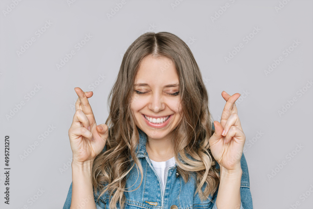 A young pretty smiling blonde woman in a denim jacket and jeans with her eyes closed crosses her fingers for good luck waiting for the results of the lottery or exams isolated on a gray background A young pretty smiling blonde woman in a denim jacket and jeans with her eyes closed crosses her fingers for good luck waiting for the results of the lottery or exams isolated on a gray background
