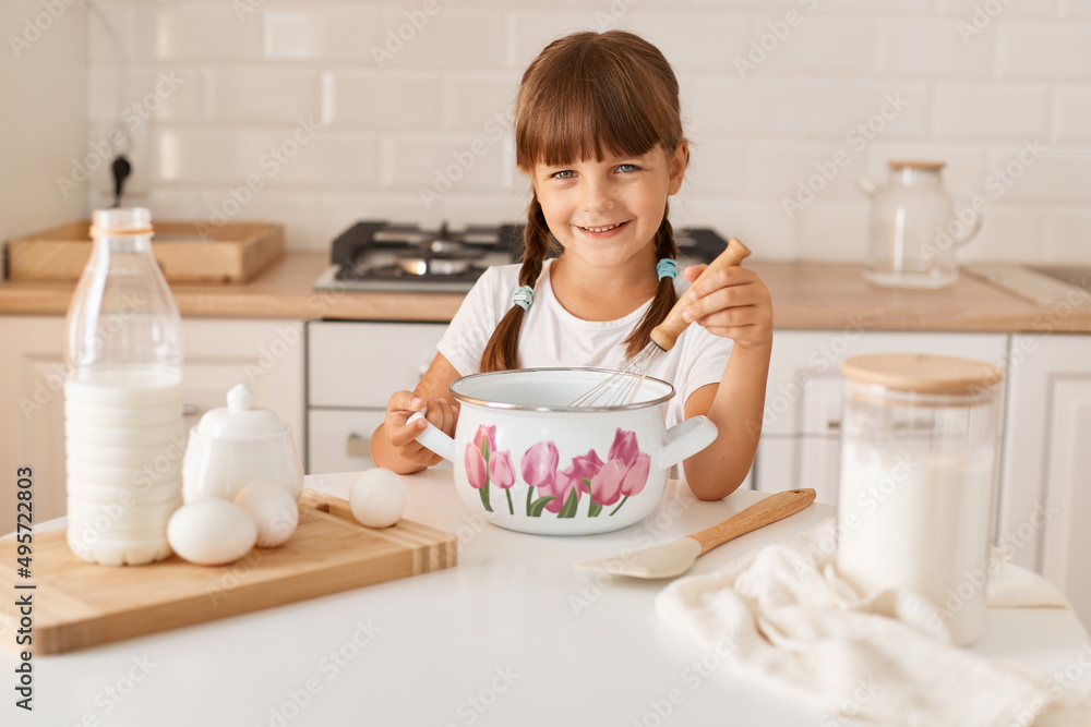 Portrait of smiling cute little girl baker with braids on kitchen with baking ingredients, looking at camera with charming smile, preparing dough for pie or cookie. Portrait of smiling cute little girl baker with braids on kitchen with baking ingredients, looking at camera with charming smile, preparing dough for pie or cookie.