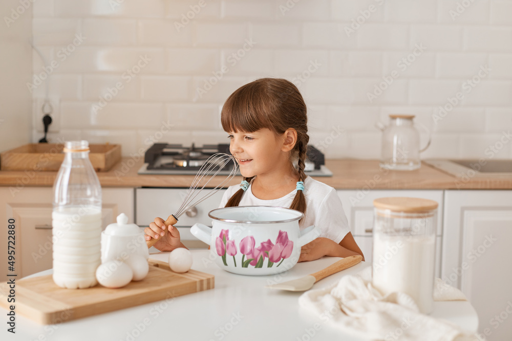 Horizontal shot of Caucasian dark haired cute little girl baker on kitchen with baking ingredients, preparing dough with milk, eggs and flour, making cake for birthday. Horizontal shot of Caucasian dark haired cute little girl baker on kitchen with baking ingredients, preparing dough with milk, eggs and flour, making cake for birthday.