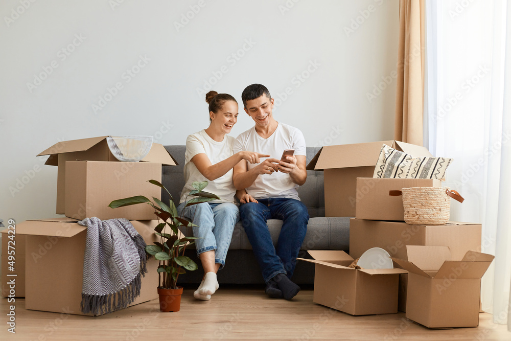 Indoor shot of positive young couple sitting on sofa surrounded with carton boxes, family during relocation, husband using phone and showing something funny. Indoor shot of positive young couple sitting on sofa surrounded with carton boxes, family during relocation, husband using phone and showing something funny.