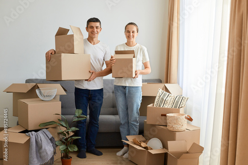 Indoor shot of satisfied positive young couple sitting on sofa surrounded with carton boxes, family during relocation to a new apartment, holding personal piles in carton parcels. © sementsova321