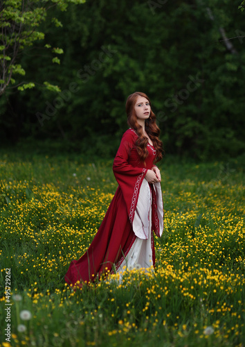 Fotografia Red haired maiden in medieval dress walking in field of yellow flowers