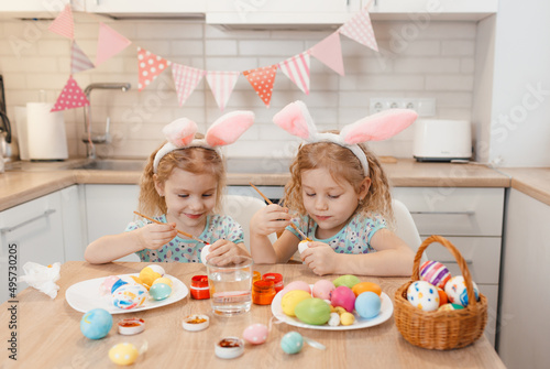 two twin girls are preparing for the Easter holiday at the kitchen table painting Easter eggs.