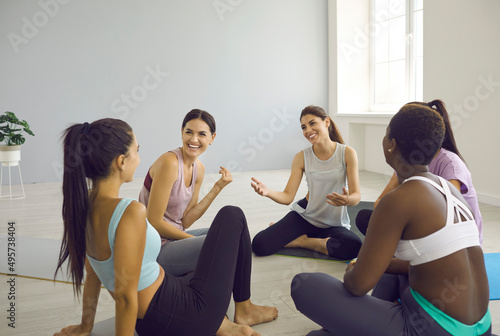 Diverse group of happy beautiful fit young women having a nice chat after a yoga class. Cheerful multiracial female friends talking and laughing sitting in a circle on yoga mats at gym or yoga studio