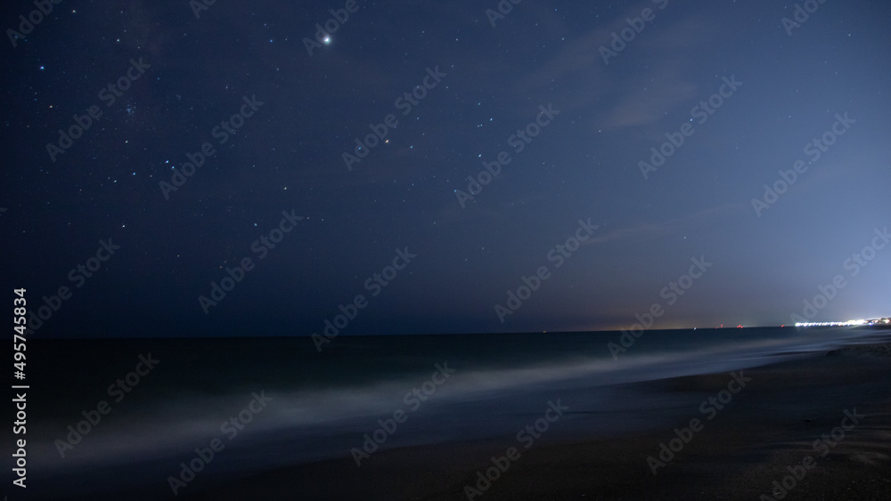 Beautiful view of a blue night sky and stars on dark background over a sandy beach and seawater