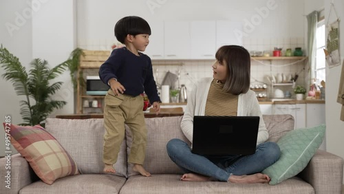 slow motion of Asian naughty preschool boy bothering his WFH mother with a laptop in living room. she shakes her head at her son while he is jumping up and down on couch