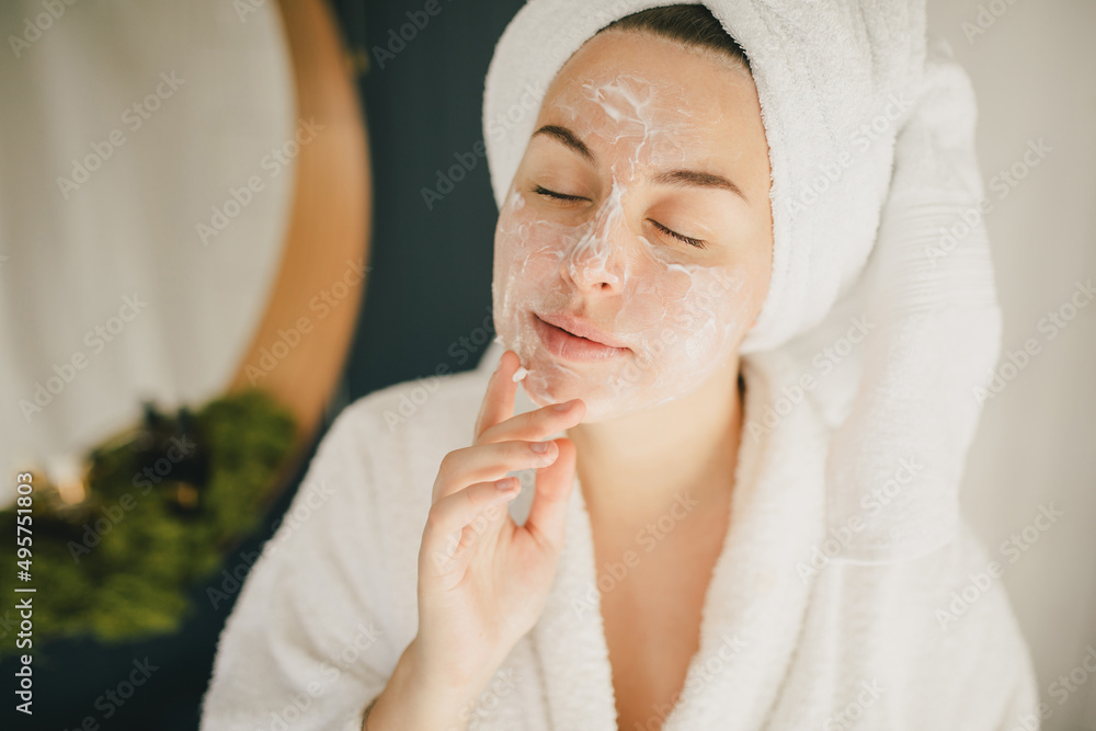 Young beautiful woman wearing bathrobe and towel on her hair applying moistrizing cream on her face. Skin care morning rituals. Beauty routine. Young beautiful woman wearing bathrobe and towel on her hair applying moistrizing cream on her face. Skin care morning rituals. Beauty routine.