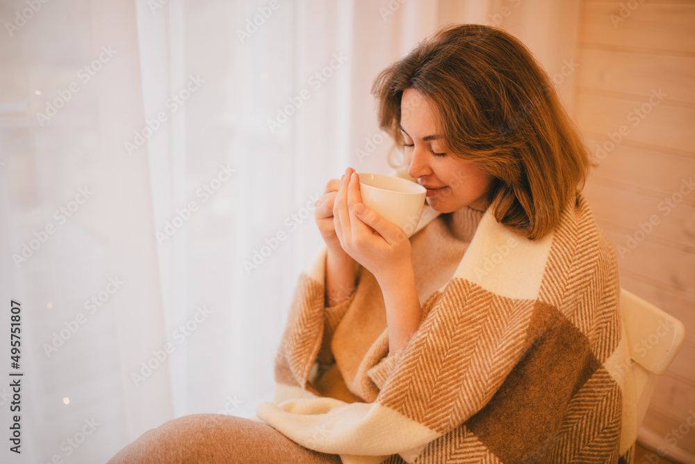 Young woman in white wrapped in blanket drinking tea near window in a cozy wooden house. Young woman in white wrapped in blanket drinking tea near window in a cozy wooden house.