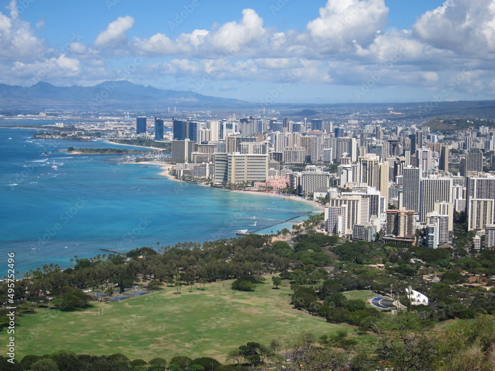 View from Diamond Head showing the Waikiki neighborhood. View of buildings, the shore, green vegetation, blue sky with clouds, blue seashore, mountains, a park with green trees. Oahu, Waikiki, Hawaii.