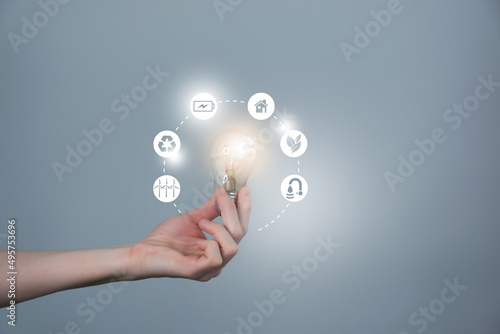 Woman`s hand holding glowing light bulb with energy resources icons. Ecological friendly and sustainable environment concept.