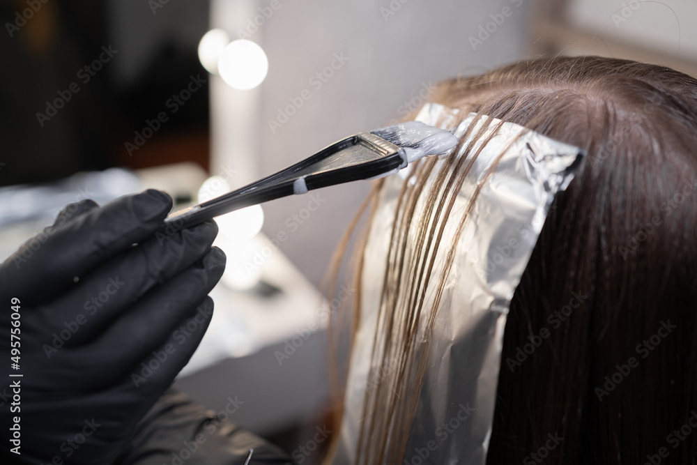Close-up of a strand of female hair on a special sheet of foil, the master applies hair dye with a brush. The process of highlighting by a professional master in a beauty salon. - obrazy, fototapety, plakaty 
