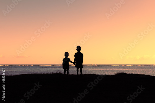 Silhouette boy and girl, brother and sister holding hands facing sunset 