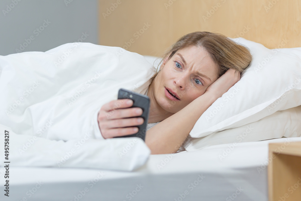 Woman lying in bed in the morning, shocked reading bad news from the phone Woman lying in bed in the morning, shocked reading bad news from the phone