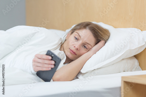 A woman lies under a blanket at home in the morning, looks at the phone screen, reads the morning news