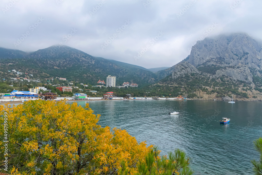 The sea bay with blue water, boats and yachts. In the foreground yellow tree foliage. In the background of the mountain and cloudy sky. Taken from the Golitsyn Trail. Novy Svet, Crimea The sea bay with blue water, boats and yachts. In the foreground yellow tree foliage. In the background of the mountain and cloudy sky. Taken from the Golitsyn Trail. Novy Svet, Crimea