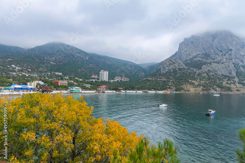 The sea bay with blue water, boats and yachts. In the foreground yellow tree foliage. In the background of the mountain and cloudy sky. Taken from the Golitsyn Trail. Novy Svet, Crimea