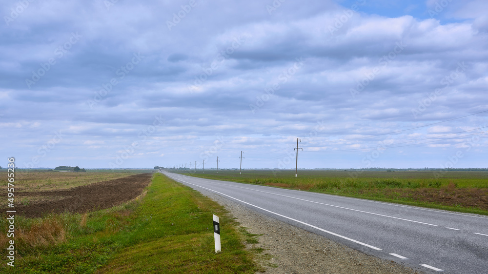 View of the asphalt road going beyond the horizon, passing by the fields. Highway with two-way traffic lanes.