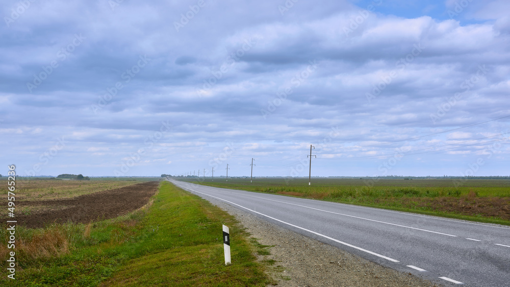 View of the asphalt road going beyond the horizon, passing by the fields. Highway with two-way traffic lanes. View of the asphalt road going beyond the horizon, passing by the fields. Highway with two-way traffic lanes.