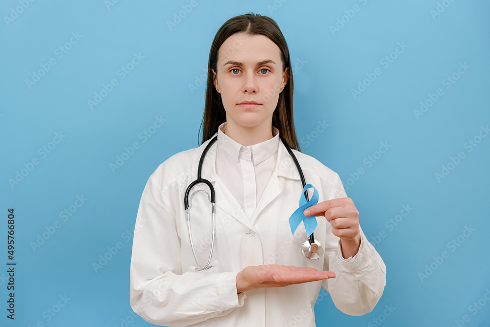 Portrait of calm young woman doctor in white medical gown with blue awareness ribbon, symbol of World Diabetes Day, looking at camera, posing isolated over studio background wall with copy space Portrait of calm young woman doctor in white medical gown with blue awareness ribbon, symbol of World Diabetes Day, looking at camera, posing isolated over studio background wall with copy space