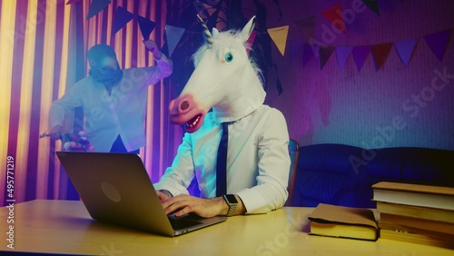 A woman in a pigeon mask is dancing in the background, and a young man in a unicorn mask is sitting at a table. Multicolored lighting. Masquerade