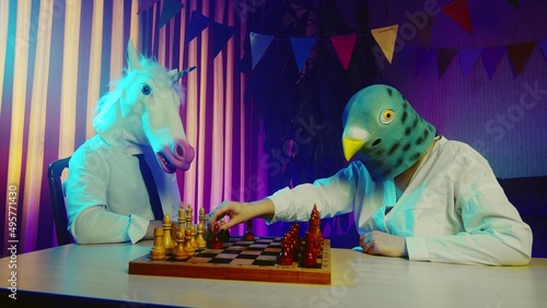A young man and a woman wearing pigeon and unicorn masks are playing chess at a table. Multicolored lighting. Masquerade