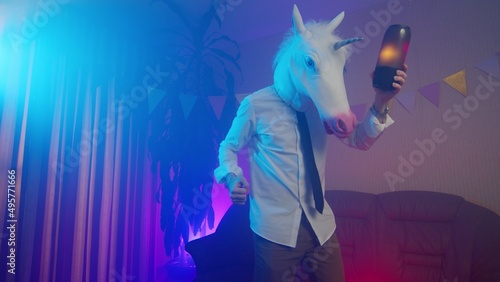 A young man in a unicorn mask is dancing with a music column in his hands. Multicolored lighting. Masquerade