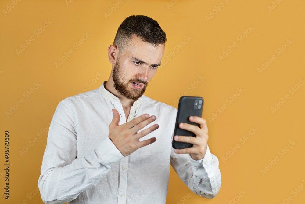 A young man on a yellow background looks at the phone and angrily talks to someone on video. Angry young man talking on the phone, negotiating. Mock up, advertising A young man on a yellow background looks at the phone and angrily talks to someone on video. Angry young man talking on the phone, negotiating. Mock up, advertising