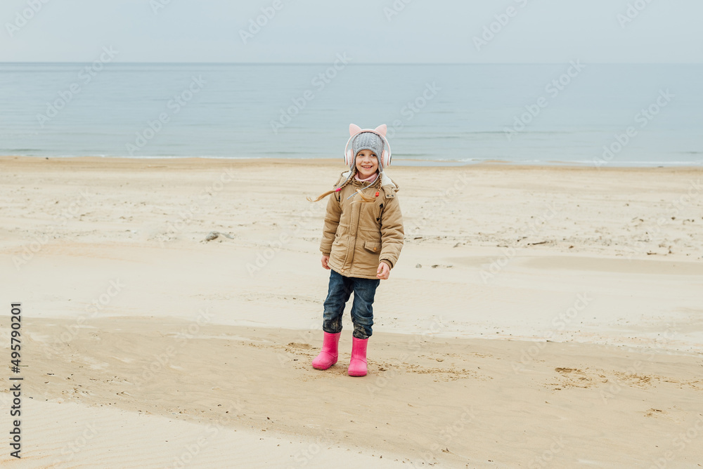 little girl listens to music in children's pink headphones with cat ears and dances on the beach the child laughs, has fun in autumn, spring on the sea coast in the off-season, non-tourist season