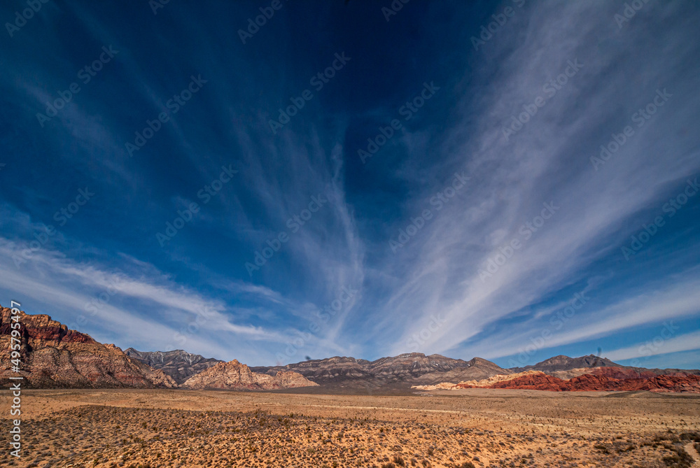 Las Vegas, Nevada, USA - February 23, 2010: Red Rock Canyon Conservation Area. Blue sky with white lines of clouds exploding out of gray mountain. Sandy flat desert floor with small scrubs.