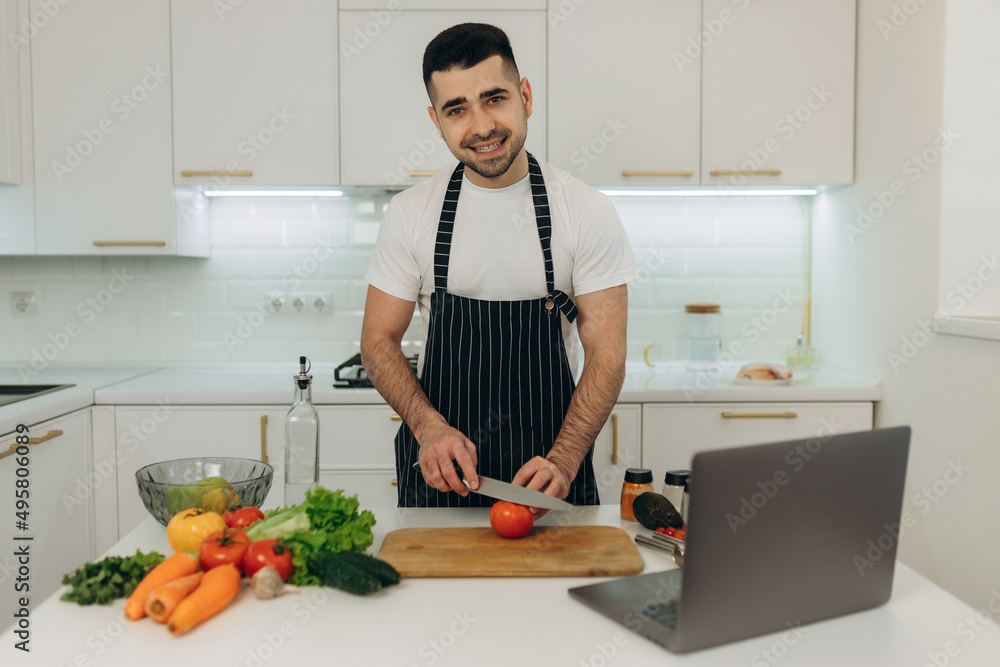 Portrait of a handsome man preparing food for himself. He looks at the laptop recipe for cooking. A man dressed in a black apron. Portrait of a handsome man preparing food for himself. He looks at the laptop recipe for cooking. A man dressed in a black apron.