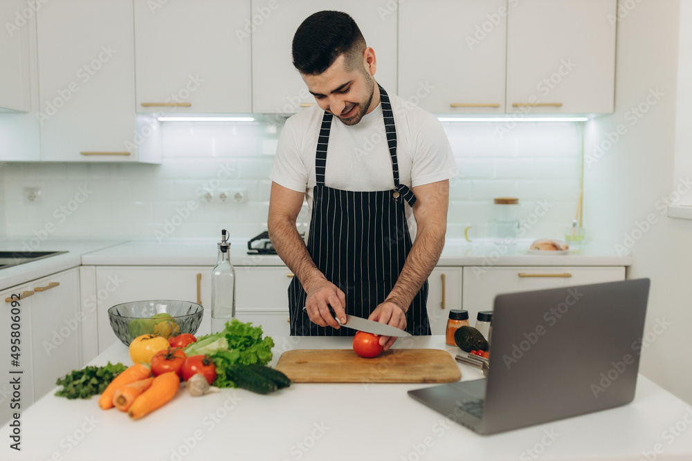 Portrait of a handsome man preparing food for himself. He looks at the laptop recipe for cooking. A man dressed in a black apron.