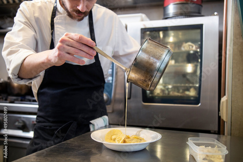 chef manos cocina cpasta fresca tortellini con salsa sirve en plato blanco en interior cocina restaurante con uniforme blanco y mandil negro close-up photo