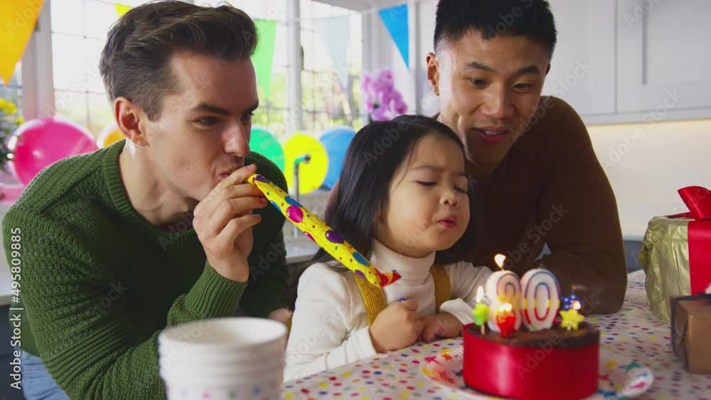 Family with two dads and daughter celebrate parent's 30th birthday with girl blowing party blower at party - shot in slow motion Family with two dads and daughter celebrate parent's 30th birthday with girl blowing party blower at party - shot in slow motion