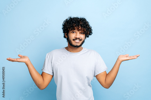 Puzzled confused indian or arabian curly-haired man in casual wear, shrug shoulders, looking questioningly at the camera, standing on isolated blue background photo