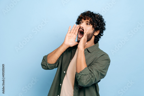 Excited indian or arabic curly haired guy in casual clothes, shouting loudly holding his hands near his mouth, making an announcement, calling out, standing on an isolated blue background