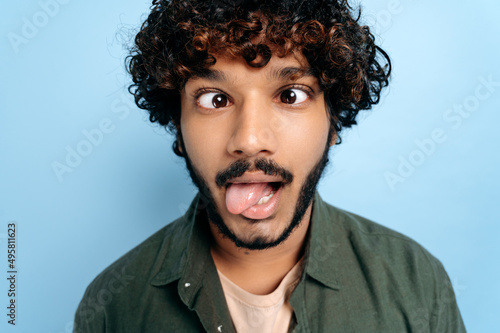 Close up face of indian or arabian curly funny guy, making a funny face, tongue out, squinted his eyes, standing on isolated blue background. Grimaces, fooling around © Kateryna