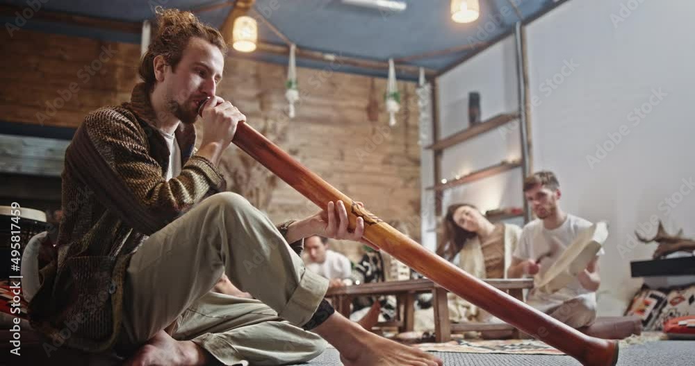 Male hippie playing music with friends. Low angle of man in casual clothes playing didgeridoo while sitting on floor and creating music with friends in loft room Male hippie playing music with friends. Low angle of man in casual clothes playing didgeridoo while sitting on floor and creating music with friends in loft room