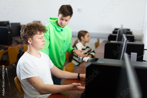 Focused teenager helping puzzled preteen schoolboy sitting at computer in college library..