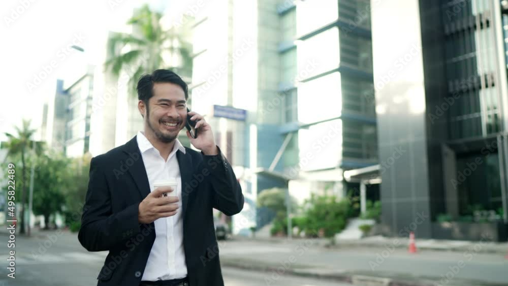 Asian businessman Smiling Wearing a Black Suit is Walking On the Streets Of the City to Work. in Hand holding a Coffee Cup using Smartphone. Urban life Style Concept. Asian businessman Smiling Wearing a Black Suit is Walking On the Streets Of the City to Work. in Hand holding a Coffee Cup using Smartphone. Urban life Style Concept.