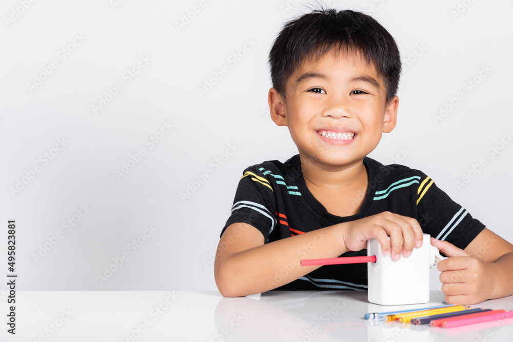 Little cute kid boy 5-6 years old smile using pencil sharpener while doing homework in studio shot isolated on white background, Asian children preschool sharpening color pencils, education concept