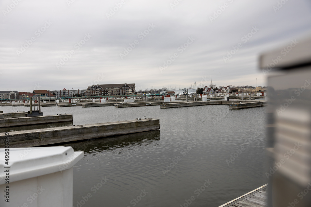 Marina boat slips empty in the fall. Wooden piers separating boat slips. Cloudy gray sky. Large multi story buildings across the harbor. White storage containers by each slip. 