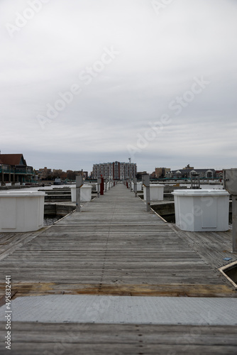 Wooden pier leading out to the water on a cloudy hazy day. Electrical posts by each boat slip. White storage containers lining the wood dock.  Large buildings seen in the background.  © Southport Images
