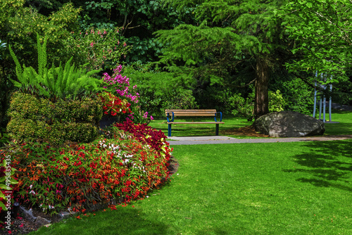Resting place at the Park in Vancouver City. Bench is located under the canopy of spreading tree on a green lawn with flower beds among flowering shrubs,   British Columbia, Canada