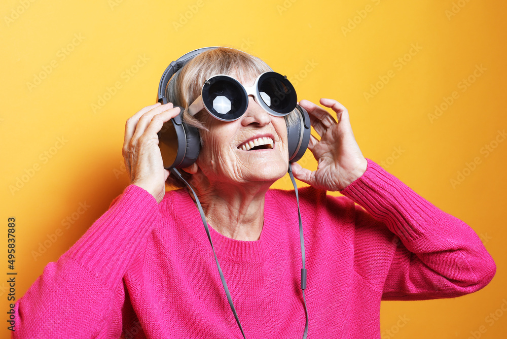 Cheerful old woman wearing pink sweater and big sunglasses listening to music with headphones over yellow background.