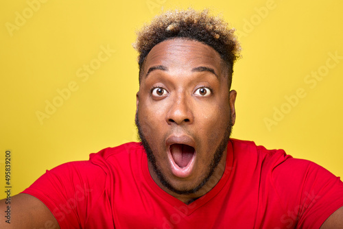 Surprised face of dark-skinned curly African-American close up in frame on yellow background. A young handsome guy opened his mouth in surprise and pulled out his eyes, pointing camera at himself © Aleksandr